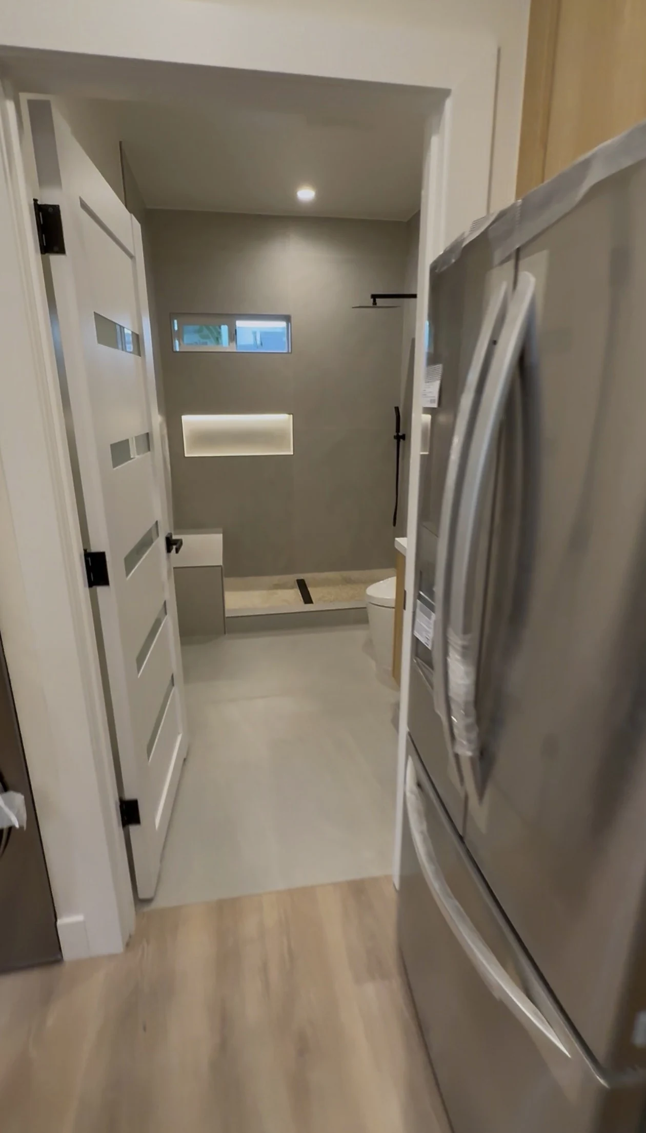 A view past the kitchen refrigerator through a frosted-panel door to the tiled shower and illuminated niche.