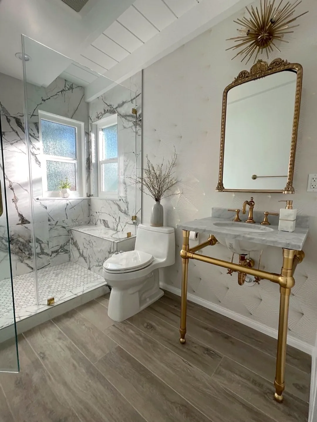 Finished Los Angeles bathroom with a dramatic white and gray marble-look shower, frameless glass, white toilet, gold-legged console sink and ornate gold mirror.