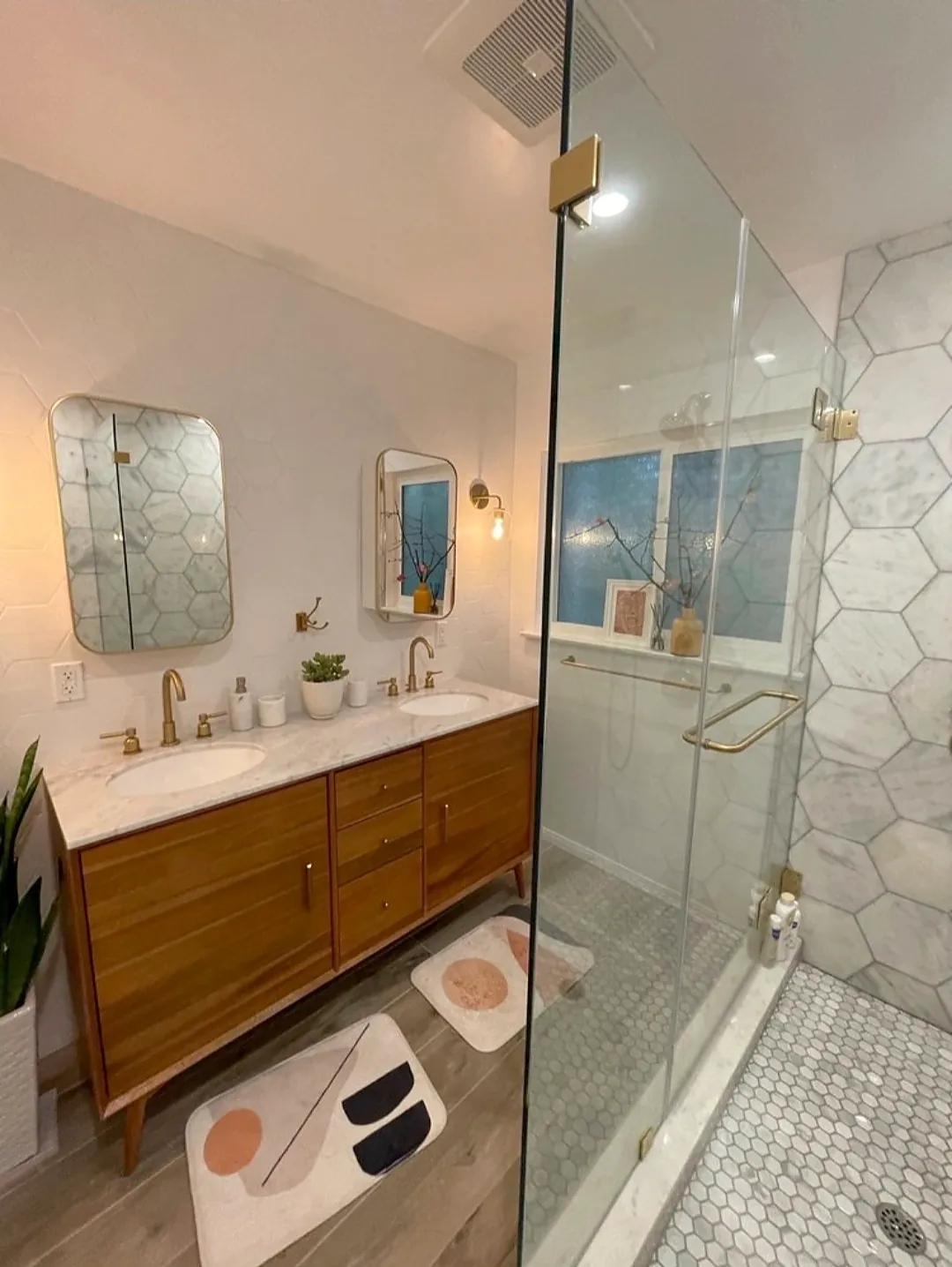 Closer angle on the wood double-sink vanity and the glass shower with marble-look hexagonal wall tile, tiny white mosaic floor and brass-tone trim.