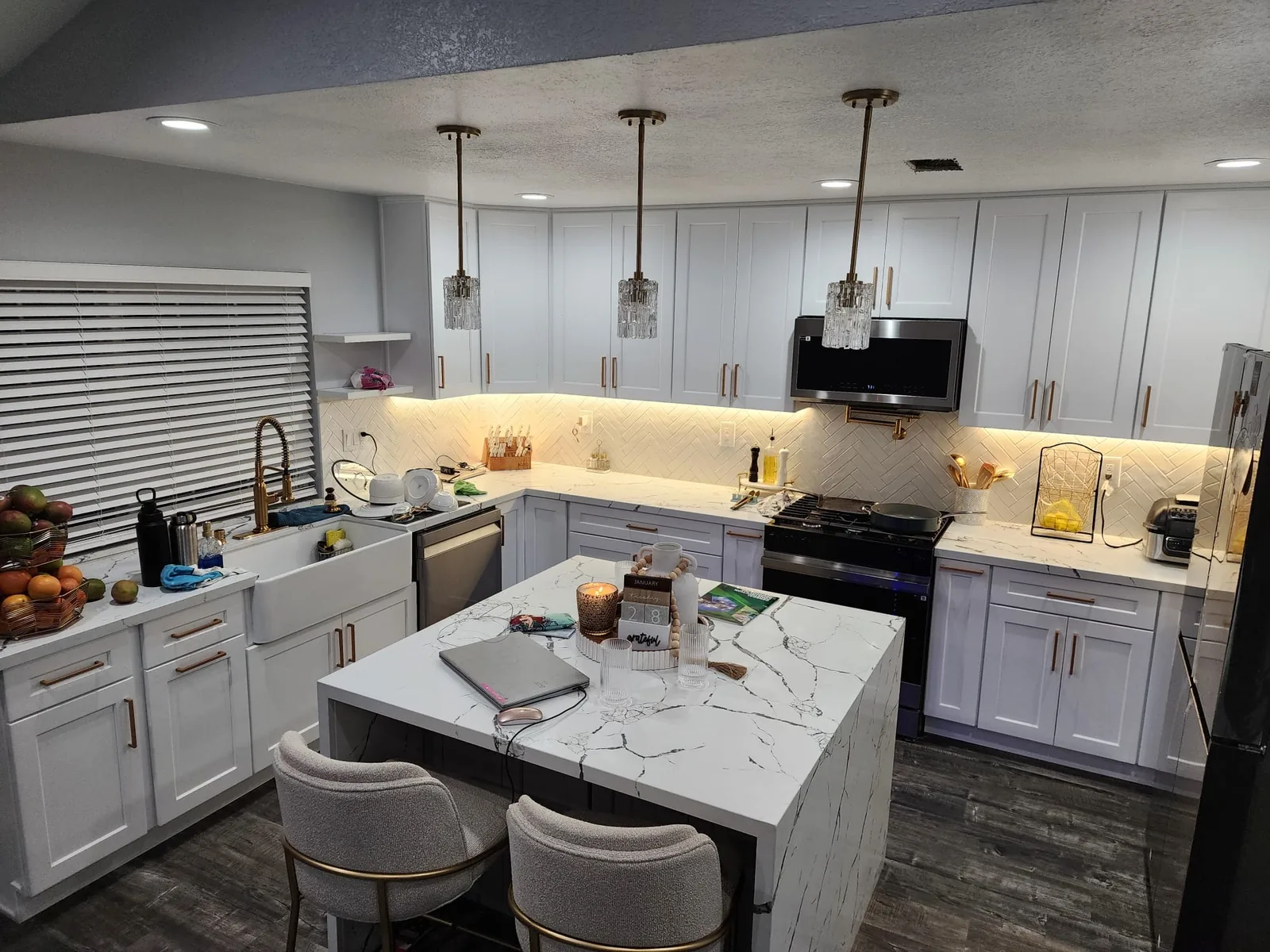 Finished kitchen with white shaker cabinets, a lit white herringbone backsplash, three glass pendants and a veined quartz waterfall island with two upholstered stools.