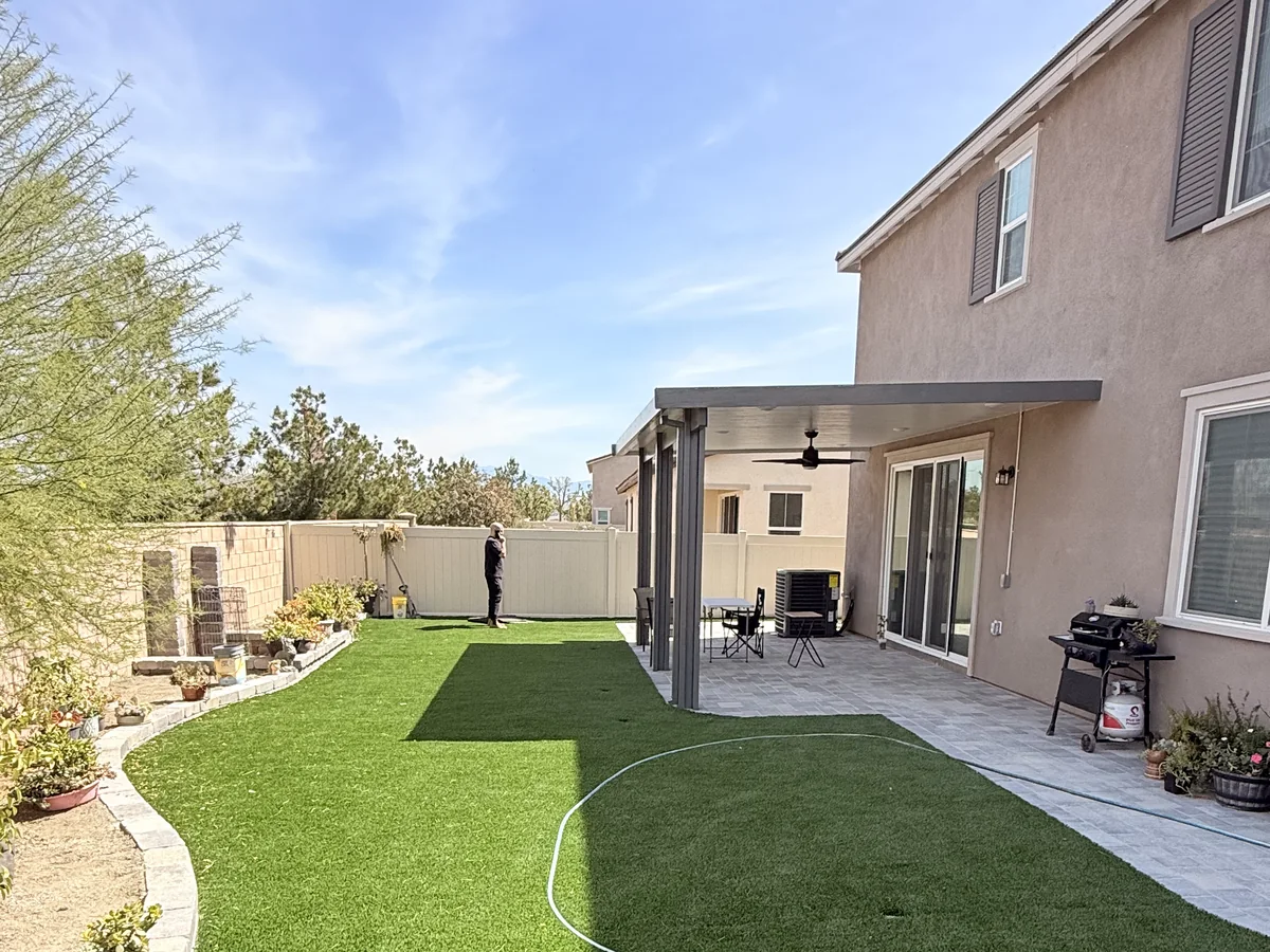 Owner-supplied yard view showing turf, a light paver patio, planting edge and gray attached patio cover beside a two-story stucco home.