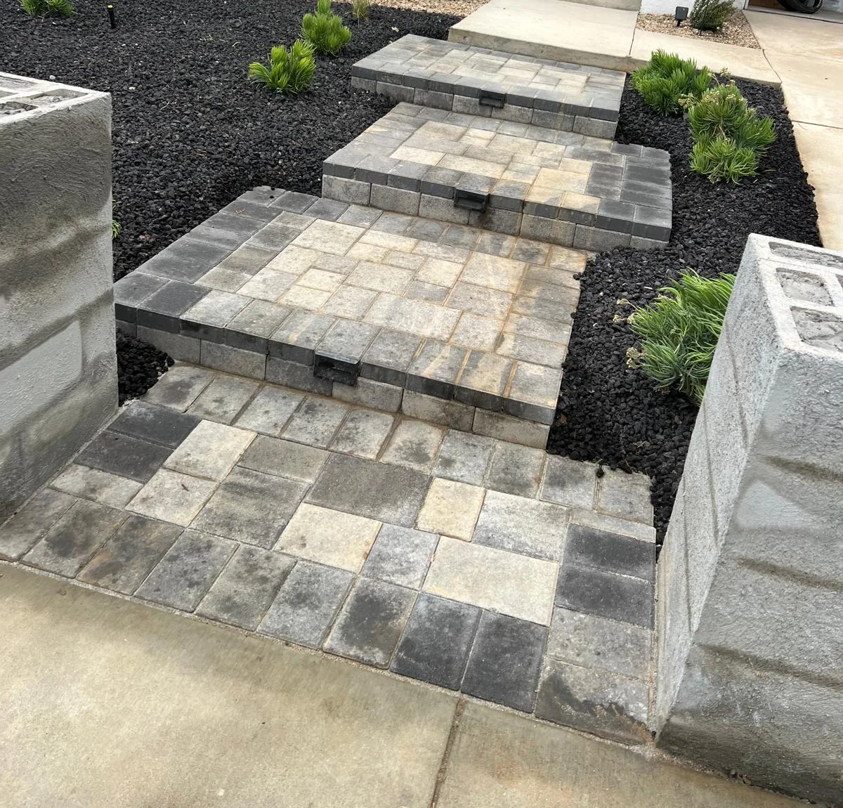 Cropped owner-supplied photo of gray paver entry steps and landings between dark decorative rock and small plants.