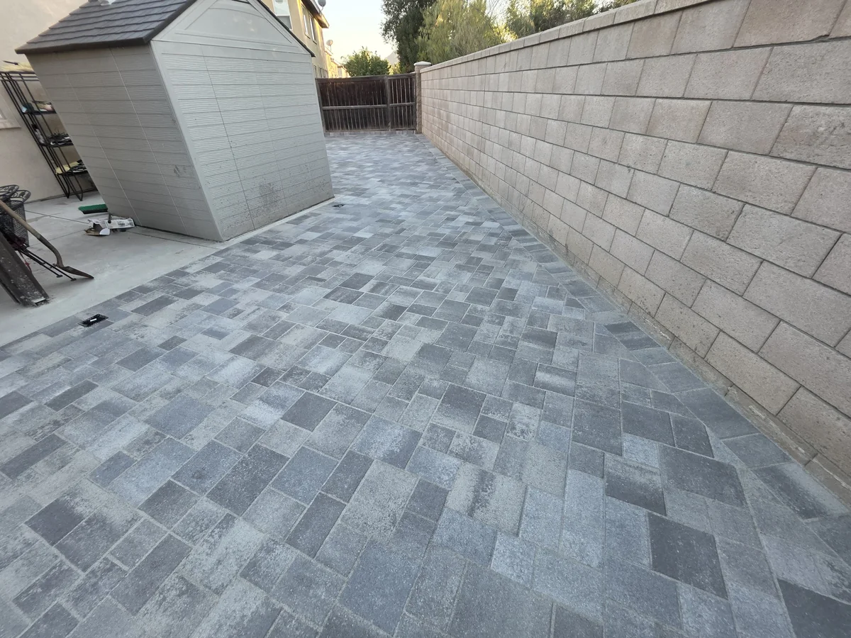 Gray mixed-size paver courtyard beside a long masonry wall and a small storage shed.