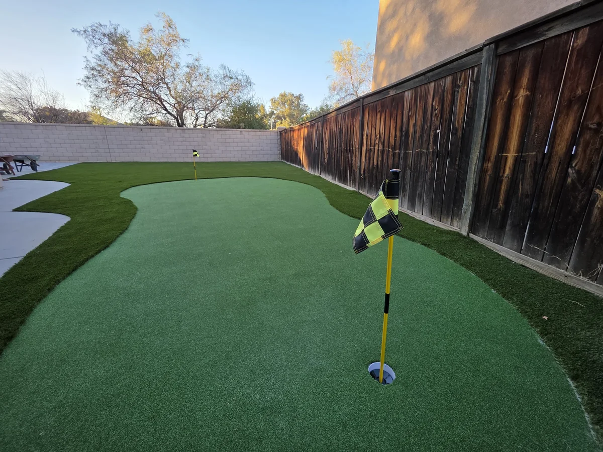 Curved putting-green-shaped turf area with a flag, light gravel and wood fence beside an ordinary stucco home.