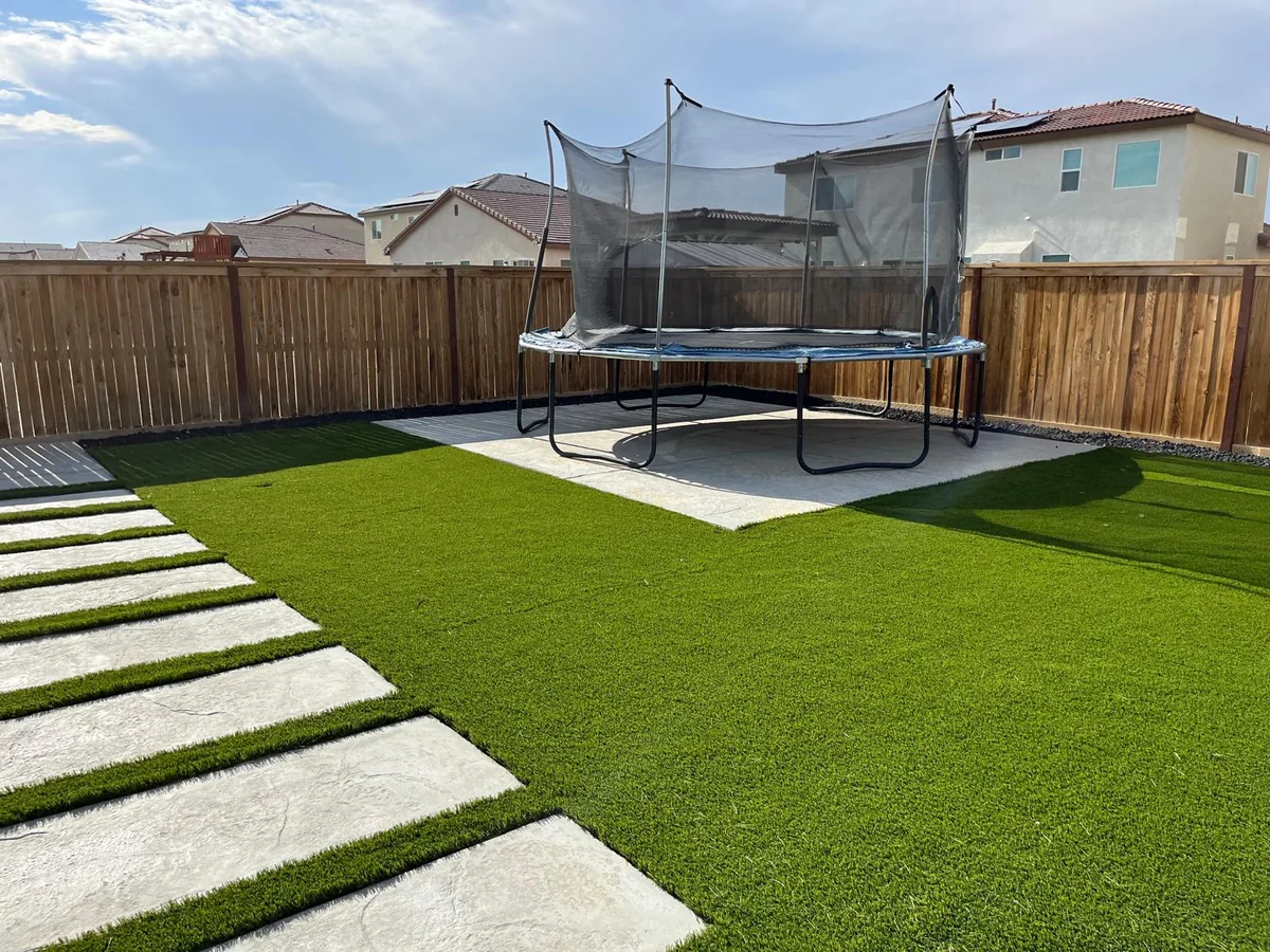 Large pale stepping stones across green turf toward a wood fence and trampoline.